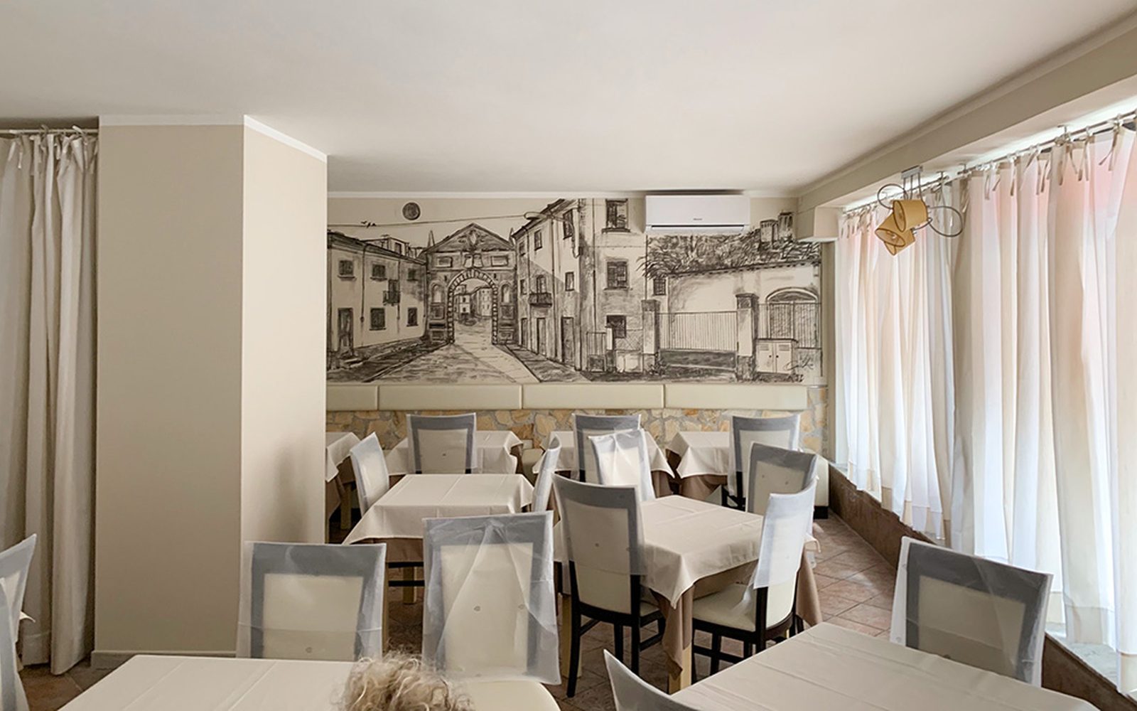 La sala del ristorante: tavoli apparecchiati di bianco davanti al grande disegno a carboncino di un arco del centro storico di Massa.